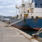 Marine service personnel reviewing a clipboard beside a merchant vessel moored at a repair quay