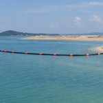 AI-generated editorial illustration showing a distant view of a floating dredging discharge pipeline approaching a reclamation shoreline