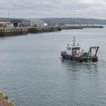 Distant stern-quarter view of a water injection dredger working in a harbour basin beside industrial quays