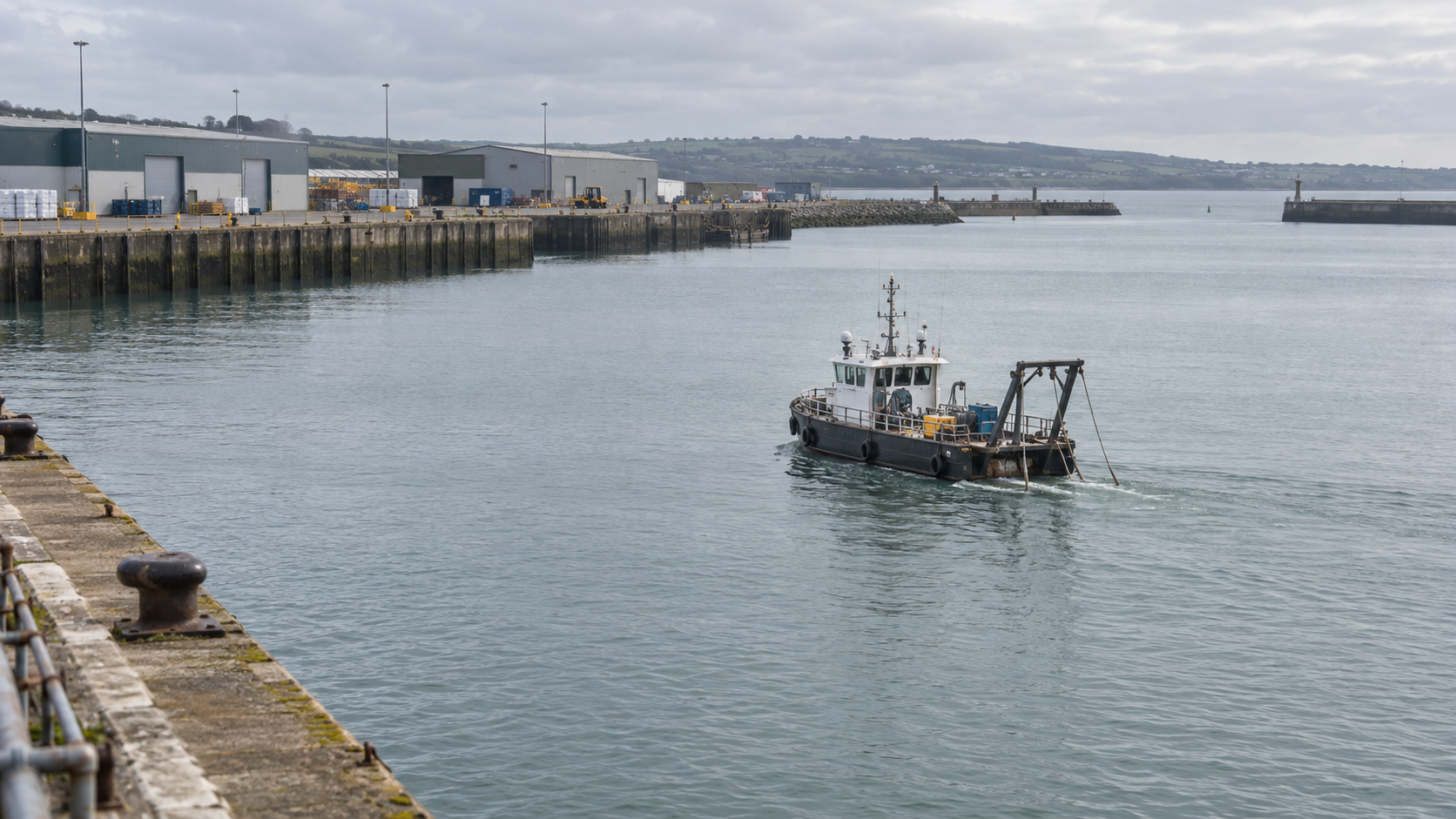 Distant stern-quarter view of a water injection dredger working in a harbour basin beside industrial quays
